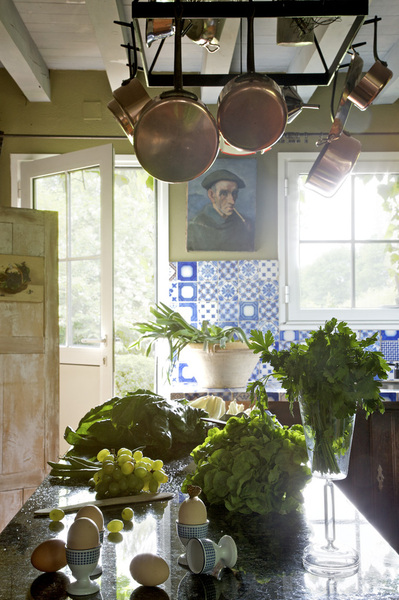 Ouverte sur le jardin,  la pièce profite des rayons du soleil tard le soir. Au-dessous d’une suspension artisanale ornée d’anciennes casseroles en cuivre, une table de drapier XIXème recouverte d’un plateau en granit plus propice à la cuisine ; peinture « Landais », Montes Iturios.