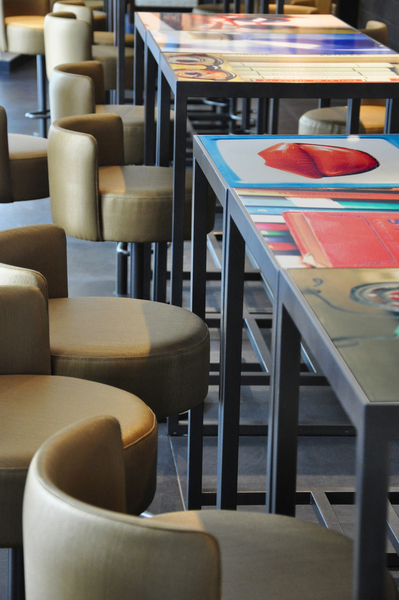 Restaurant Velvet. Gros plan sur les tables à hauteur de comptoir bar, les plateaux des tables ont été réalisés sur mesure en stratifié avec inclusion d’une photo illustrant le thème de l’univers d’un artiste fil conducteur de la décoration de l’hôtel. Les tabourets de bar ont été dessinés spécialement pour le projet.