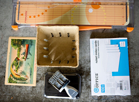 Le matériel nécessaire se compose d'une cagette à fruits, de fiches cartonnées, d'un tampon dateur, de cartes postales et d'un massicot.