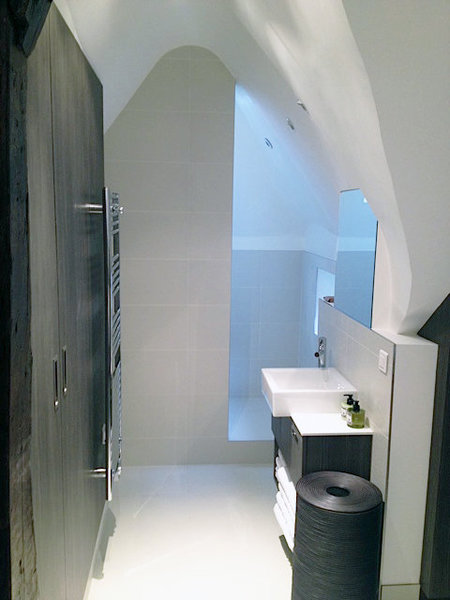 En continuité de la chambre, une salle de bains discrète a été aménagée. La douche aménagée dans une alcôve semi-ouverte, est partiellement cachée par un pan de mur carrelé, et les rangements offrent une unité esthétique. Au sol, les deux espaces se différencient.