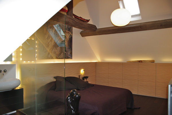 Délimitée par un mur en verre dépoli, la salle de bains ouverte fait partie intégrante de la chambre. Les couleurs chaudes et les lumières intégrées à la tête de lit qui se prolonge en rangement sous la pente de toit participent à l’ambiance tamisée et douillette de cette pièce.