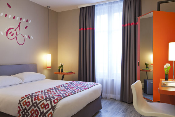 Pour la nouvelle formule d’Ibis, Ibis Style, les chambres ont été conçues dans un esprit pop et coloré. Sol en PVC façon parquet de chez Amtico, plaid créé sur mesure par Anne-Catherine Pierrey, et fabriqué par Estelle Scheibel, luminaires créés spécialement par Matec. L’orange flashy de l’armoire pivotante miroitée se retrouve comme une ponctuation en galon sur les rideaux et les abat-jour, ainsi que sur le plateau des chevets et de la table servant de bureau  (Hôtel Ibis Style, Dijon).