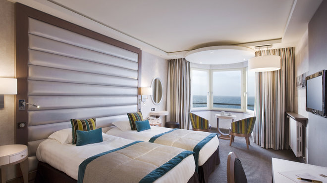 Une chambre avec une fenêtre avancée sur la mer comme un bow window, donne l’impression d’être sur un bateau de croisière. Les dessus de lit et les coussins en velours bleu canard apportent une touche d’élégance dans un esprit grand large. (Le Grand Hôtel des Thermes, Saint-Malo).