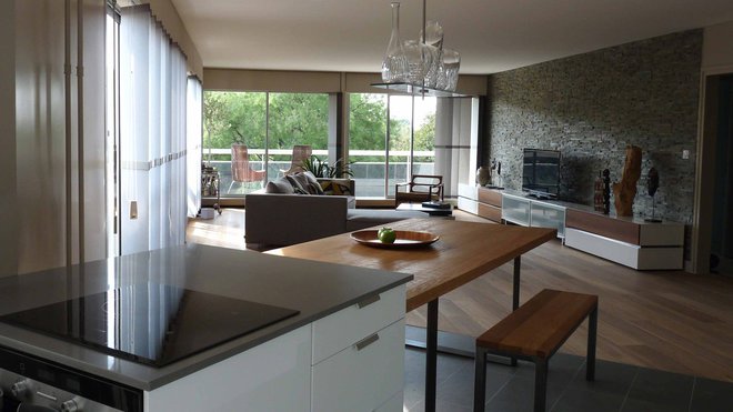 Les baies vitrées sont protégées par des stores à lamelles qui domptent la lumière, sans cacher le décor naturel  extérieur. Une table en bois sur mesure est surmontée d’un lustre (création Philippe Starck pour Baccarat), qui apporte une touche de raffinement à ce coin repas accolé à l’îlot central de la cuisine.