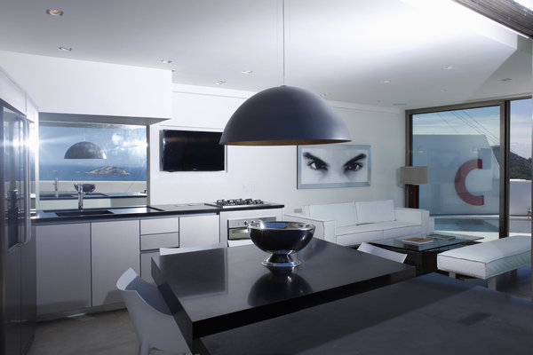 La cuisine graphique est meublée d’une grande table en granit noir, conçue en deux parties. Un second plateau permet de prendre ses repas à l’extérieur. Un salon immaculé s’ouvre sur la piscine. Suspension lumineuse avec intérieur en métal doré à la feuille, Via Manzoni.