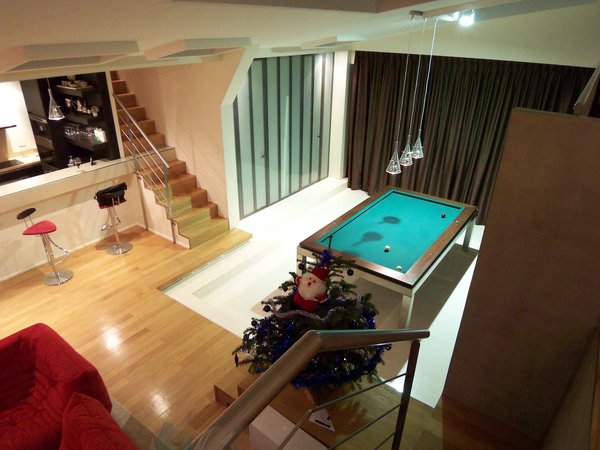 Le billard est encadré par un emmarchement, et repose sur un sol en résine qui contraste avec le plancher. La porte en acier et verre opalescent, d’esprit industriel, mène à la buanderie, à l’arrière cuisine et aux toilettes.