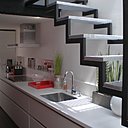 Appart 2. L’escalier aérien qui permet d’accéder au 3ème niveau s’accroche au plafond de la cuisine, avec ses éléments en enfilade.