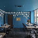La salle de restaurant, à mi-chemin entre tradition et modernité, est mise en valeur par un parquet massif de couleur sombre, et une peinture bleu turquoise. Des assiettes de porcelaine Bernardaud créent une décoration murale unique et originale, qui contraste avec la simplicité du mobilier, tables de bistrot et chaises « Surpil » chez Gras.