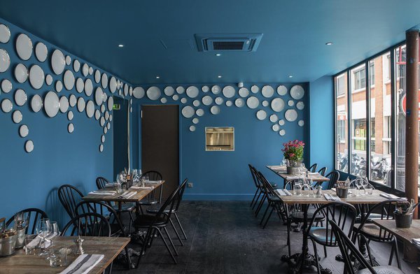 La salle de restaurant, à mi-chemin entre tradition et modernité, est mise en valeur par un parquet massif de couleur sombre, et une peinture bleu turquoise. Des assiettes de porcelaine Bernardaud créent une décoration murale unique et originale, qui contraste avec la simplicité du mobilier, tables de bistrot et chaises « Surpil » chez Gras.