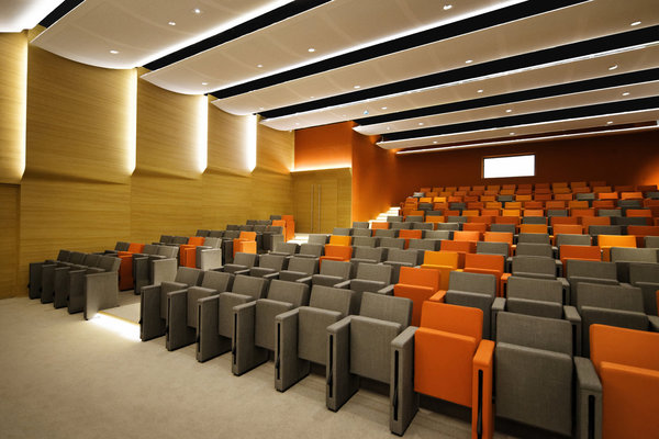 Les parties basses des murs de l’auditorium sont tapissées de panneaux en chêne micro-perforé. Au plafond, du staff perforé réfléchissant. Les courbes des cloisons et les taches de couleurs des fauteuils contrastent sur le béton blanc du bâtiment extérieur historique.