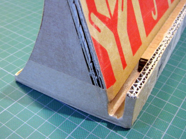 Etape 8 : Découpez des morceaux de papier kraft gommé. Humidifiez la face brillante en passant le morceau de kraft sur l’éponge humide. Recouvrez soigneusement votre construction, au niveau de chaque découpe, de chaque jonction pour avoir des côtés bien uniformes et masquer ainsi les inscriptions non désirées.