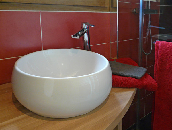 Dans la salle de bains des chambres d’hôtes, on retrouve le même code couleur, avec un carrelage rutilant qui s’associe à la chaleur du bois et la pureté immaculée d’une vasque en céramique à la forme organique.