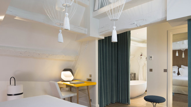 Sous les toits, la chambre Clara Gazul affirme un charme authentique avec un coin bureau et une douce séparation entre la chambre et la salle de bains. Rideau en velours de coton, Lelièvre. Suspension de Matteo Ugolini, Karman.