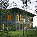 A la tombée de la nuit, la maison est éclairée grâce aux façades vitrées. Le toit à corniche fait partie de l’esthétique de l’architecture et intègre aussi la technologie : panneaux solaires, éolienne, végétaux…