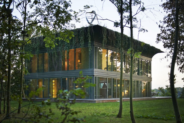 A la tombée de la nuit, la maison est éclairée grâce aux façades vitrées. Le toit à corniche fait partie de l’esthétique de l’architecture et intègre aussi la technologie : panneaux solaires, éolienne, végétaux…