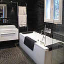 Les murs de la salle de bain sont recouverts de carrelage noir pour contraster avec la baignoire et les équipements sanitaires immaculés. Ils s’opposent avec l’autre côté de la pièce carrelé en blanc.