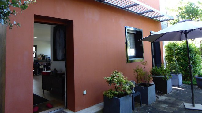 La maison en béton est recouverte d’un enduit couleur rouge argile, qui contraste avec les brise-soleils métalliques et les pots des plantations couleur zinc.