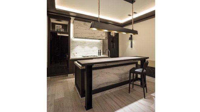 L’implantation en îlot central permet de valoriser le plan de travail en marbre, le bar en bois massif. Les sources d’éclairage indirect accentuent l’ambiance feutrée et intimiste.