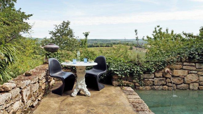 La piscine dotée d’une fontaine est encadrée par des margelles en pierre brute. Table guéridon en porcelaine revisitant le style des meubles d’extérieur en ciment, avec des formes de tronc et branches d’arbre très en vogue au milieu du XXème siècle. Chaises en polypropylène, modèle Panton Chair édité par Vitra.