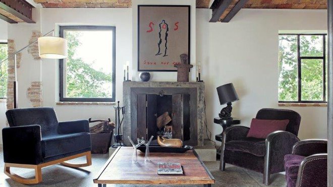 Comme dans une maison traditionnelle, le salon s’organise autour de la cheminée en tadelakt au foyer occulté par des portes en métal. A gauche, les deux fauteuils aux accoudoirs cloutés proviennent du grand-père de Marc Hertrich qui possédait une fabrique de meubles. A droite, le fauteuil à bascule est une création du studio MHNA et typique de son style, rencontre de deux styles, créé pour un hôtel. Au–dessus de la cheminée, une sérigraphie de Bernard Quesniaux.