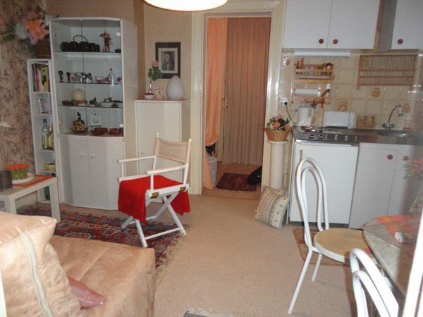 La cuisine avant : une pièce de vie maladroitement agencée avec un coin cuisine sans plan de travail implanté comme une verrue. Une décoration chargée, un espace encombré et trop meublé.