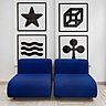 Devant 4 sérigraphies (série Simboli sinsemantici) d’Enzo Mari, 2 fauteuils en mousse houssés avec piètement en acier chromé (L 75 x L 86 x H 66 cm). Existe en bleu de France  ou noir. Collection Suzanne Lounge, création Kazuhide Takahama éditée en 1965 par Dino Gavina.