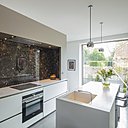 La cuisine design et graphique est dotée d’un îlot central. Les meubles blancs monolithiques contrastent avec les couleurs sombres et minérales de la crédence.