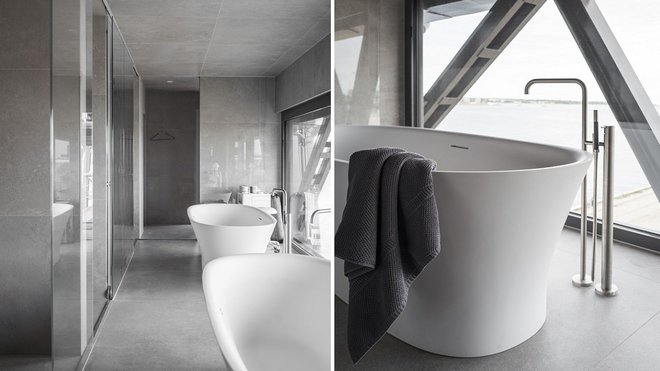 Nappée d’un dallage de pierre naturelle, la salle de bains est dotée d’une baignoire en îlot, se reflétant dans un miroir, Elle offre un mur entièrement vitré sur le port.