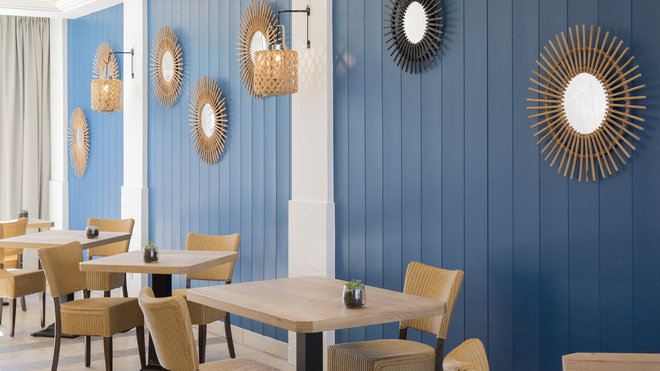 Dans le restaurant, la décoratrice a réutilisé les éléments décoratifs et les chaises du lieu d’origine en rotin et osier qu’elle a mis en valeur sur la toile de fond d’un lambris mural bleu. Dans le restaurant, la décoratrice a réutilisé les éléments décoratifs et les chaises du lieu d’origine en rotin et osier qu’elle a mis en valeur sur la toile de fond d’un lambris mural bleu.