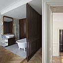 La baignoire de la salle de bains en cristallin avec vasque assortie (modèle Ottoccento, chez Agape) est occultée par un claustra en noyer d’Amérique. Elle est composée de lames de bois verticales à claire-voie implantées de façon oblique qui dessinent une sorte de paravent. L’intimité du lieu est sauvegardée tout comme sa luminosité. Robinetterie, création Patricia Urquiola chez Axor. Appliques lumineuses, modèle Métro 22, chez Décor Walther.