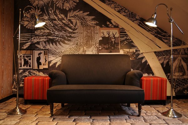 Sous les toits, un effet de dentelle XXL et des pages de carnets de tendances animent le mur qui met en valeur un canapé monochrome posé sur un sol animé d’un effet de trompe-l’œil de pavement ancien. Les lampadaires de type industriel et les poufs carrés apportent une touche plus actuelle.