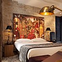 En tête de lit, sur un mur de pierre apparente, bâche imprimée d’un tableau avec détails de carnets de croquis, études et recherches de Christian Lacroix. En tête de lit, sur un mur de pierre apparente, bâche imprimée d’un tableau avec détails de carnets de croquis, études et recherches de Christian Lacroix.