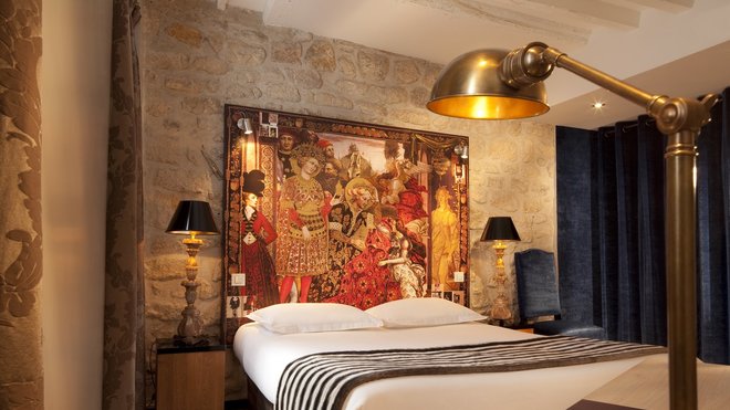 En tête de lit, sur un mur de pierre apparente, bâche imprimée d’un tableau avec détails de carnets de croquis, études et recherches de Christian Lacroix. En tête de lit, sur un mur de pierre apparente, bâche imprimée d’un tableau avec détails de carnets de croquis, études et recherches de Christian Lacroix.