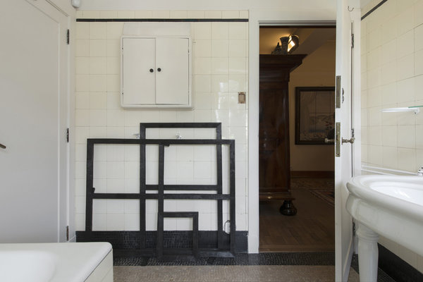 Dans la salle de bains, en contraste noir sur le carrelage blanc, cadres en bois sombre qui accentuent le côté graphique et bicolore de l’espace, au sol marqué par une mosaïque avec bordure et plinthe noires. Œuvre « If things are not as you wish them as they are » de Stefan Nikolaev.