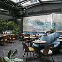 La salle de restaurant sous sa charpente métallique vitrée offre des allures de jardin d’hiver. Chaises Tatra vin­tage, pla­teau de tables en bois debout, piè­te­ments en béton, cana­pés recou­vert d’un velours lavé, rideaux en daim…