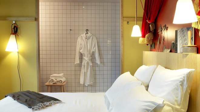 La salle de bains minimaliste en carrelage blanc contraste sur les murs jaunes. Une barre d’appui en bois permet d’accrocher ses affaires, un luminaire… Ici lampe baladeuse à poser ou suspendre, création Konstantin Grcic, modèle May Day chez Flos.